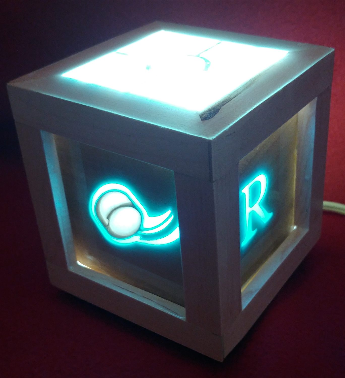 A wooden cube lamp glowing teal, its panels etched with a hockey logo and a monogram letter.